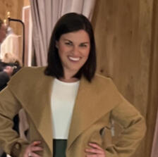 woman poses with a big smile on her face in her new clothes, wearing a camel colored wide lapel collared coat and white form fitting t-shirt